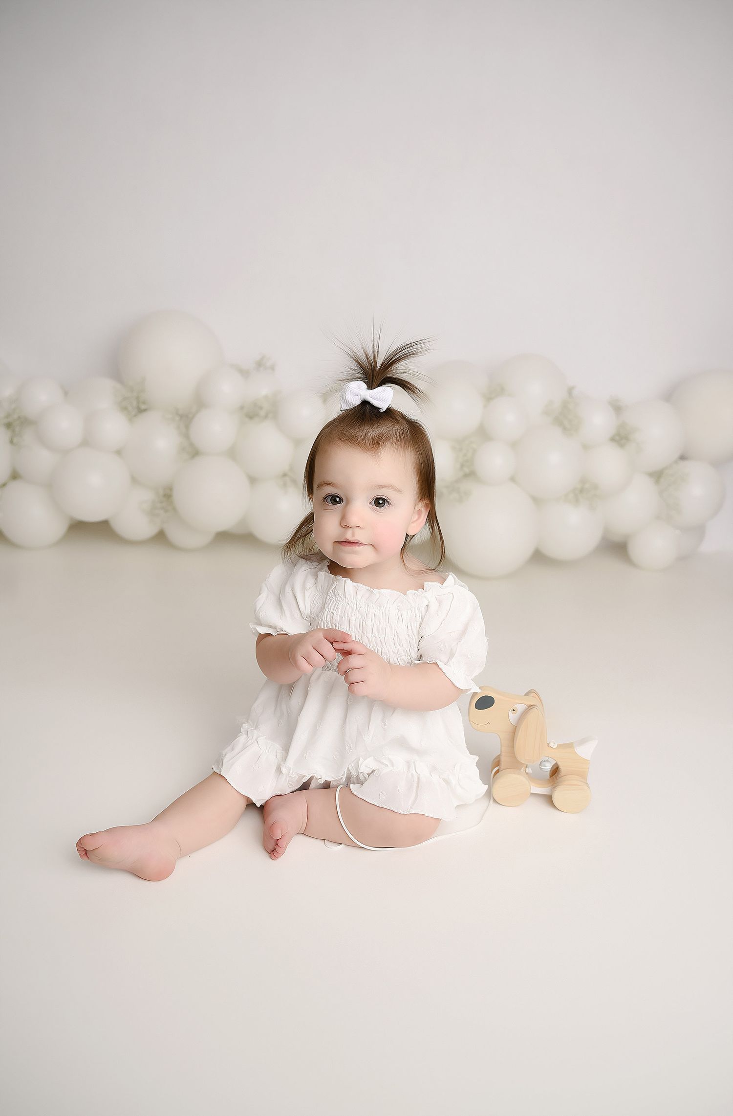Cute baby girl in white romper sits with wooden toy dog against white balloon wall backdrop.