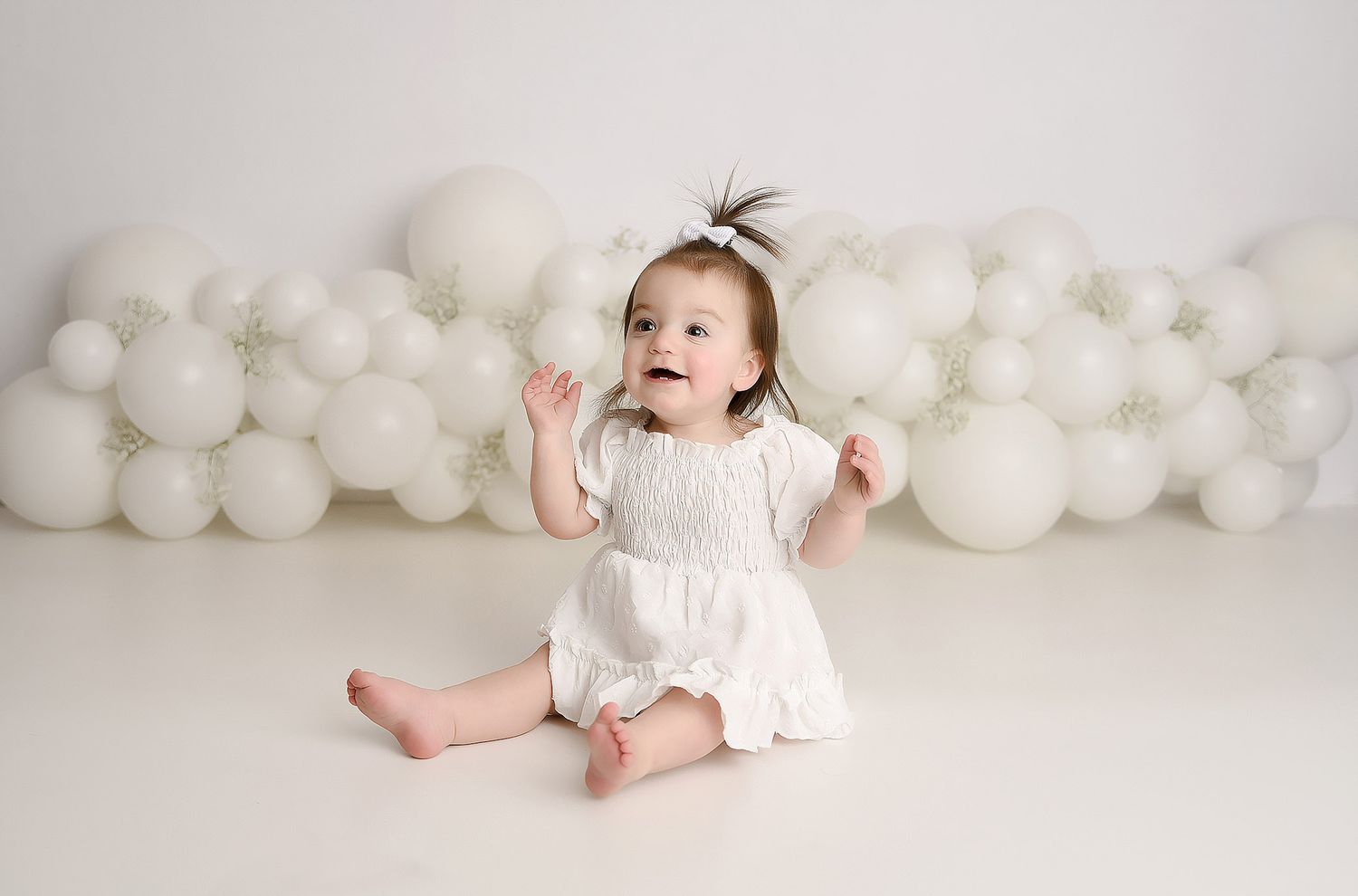 Happy baby girl in white romper sits barefoot against white balloon wall backdrop looking up.