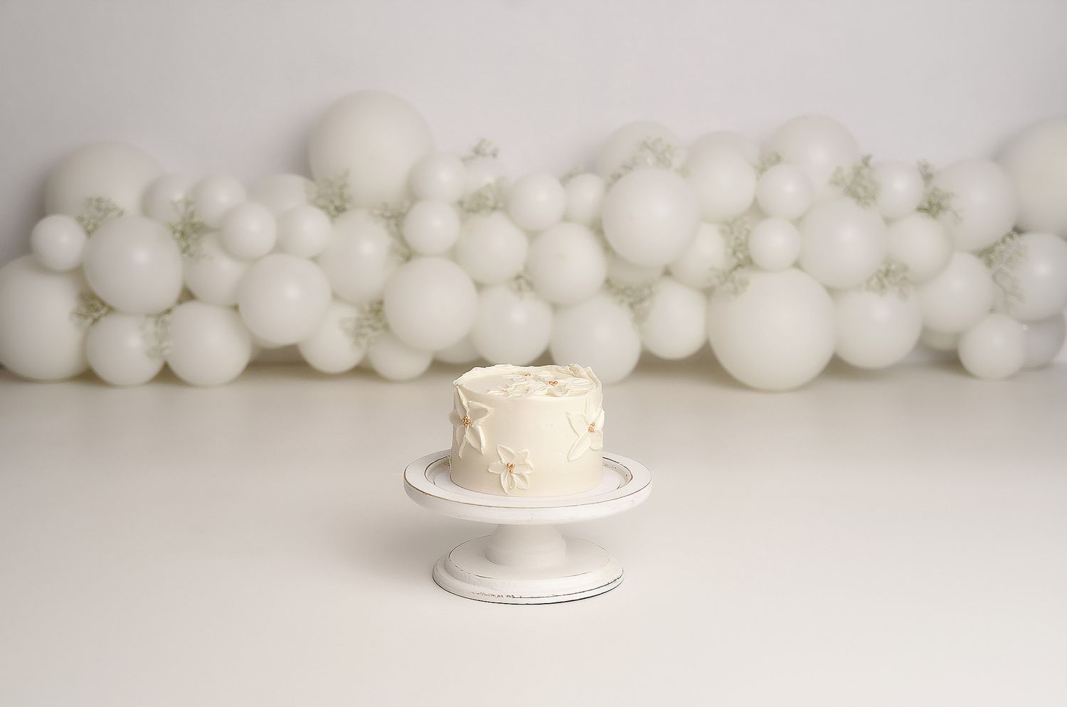 Simple white frosted smash cake on white pedestal stand against all-white balloon wall backdrop.