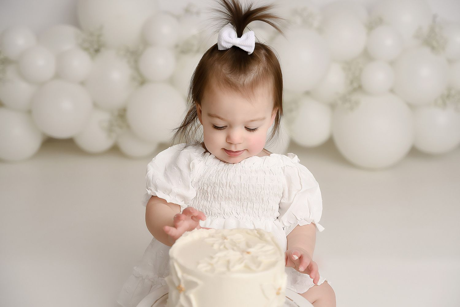 Baby girl in white dress touching her first birthday cake at a white balloon backdrop cake smash photo session.