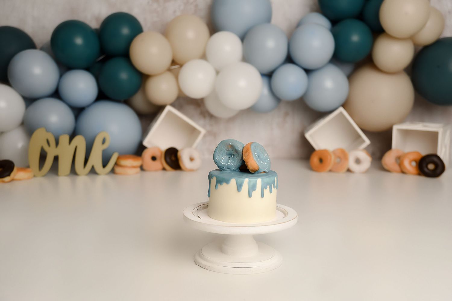 Blue drip smash cake on white stand with balloon garland and donut decorations for first birthday.