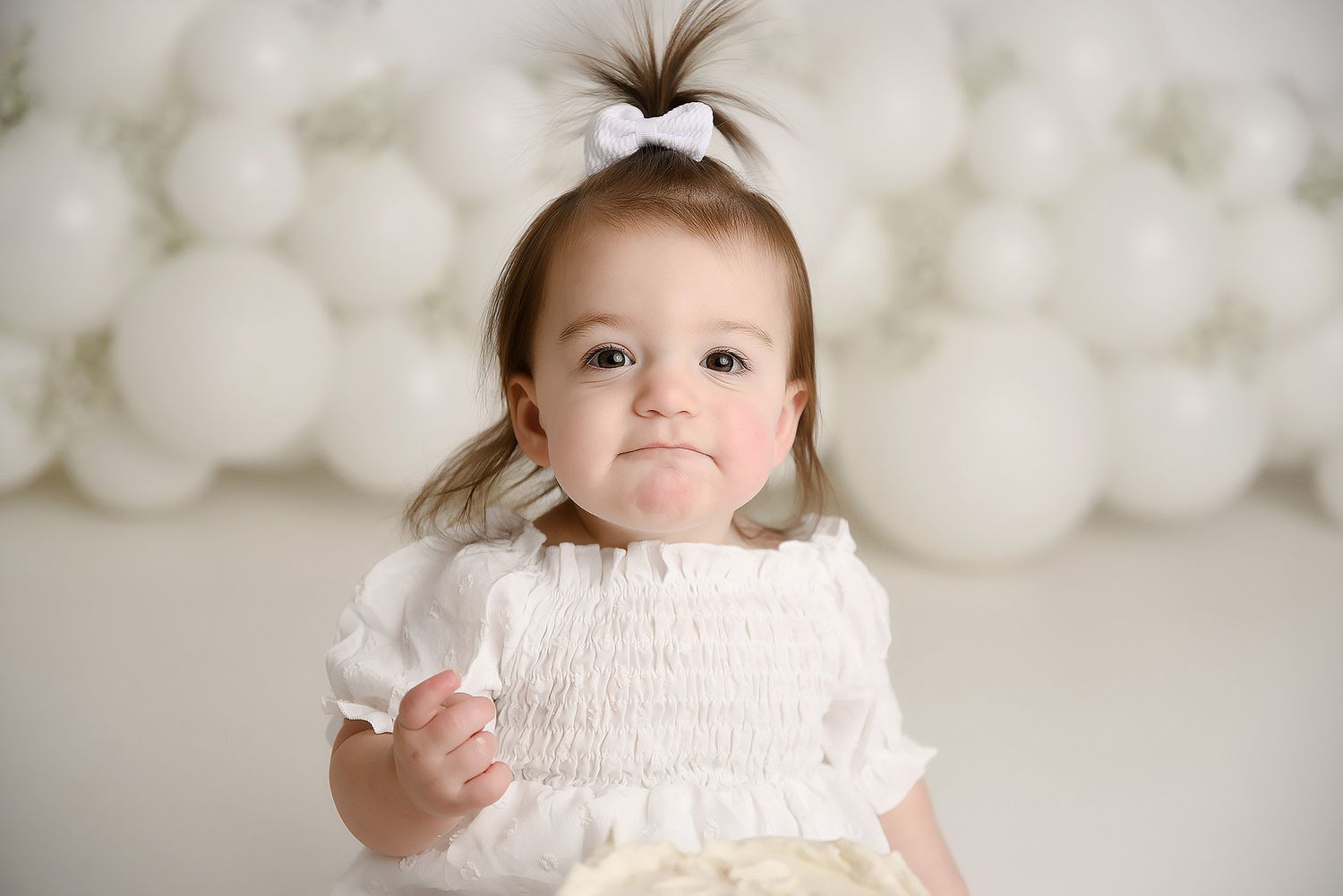 Adorable baby girl in white smocked dress with bow hair tie posing at a white balloon backdrop photo session.