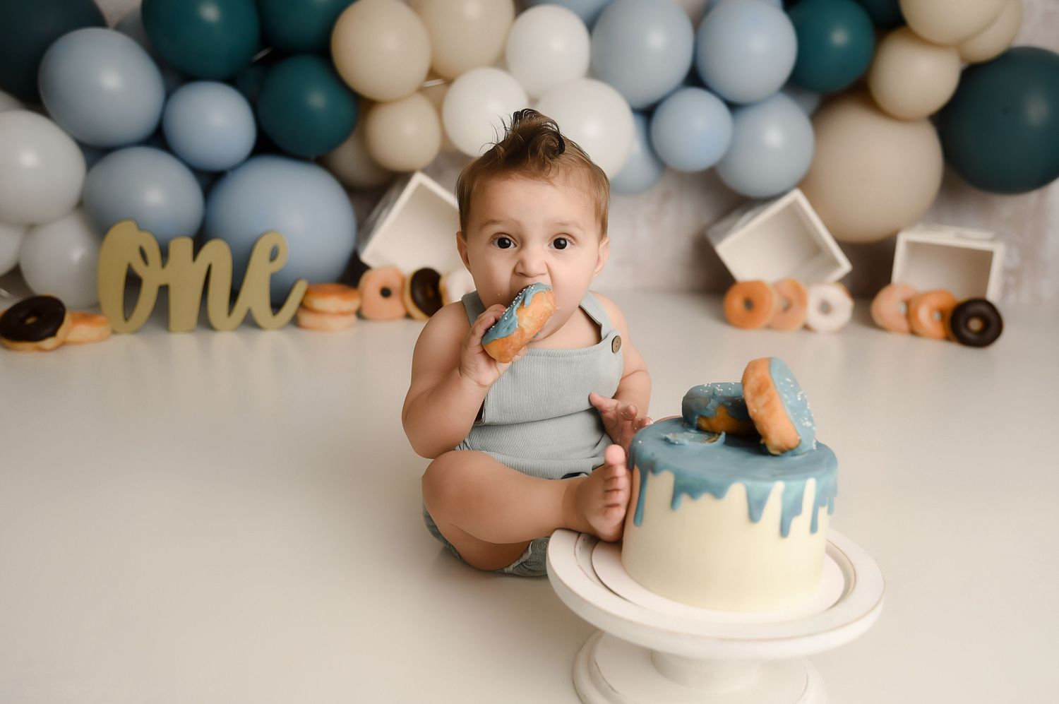 Baby boy eating donut at first birthday cake smash with blue balloons and one sign.