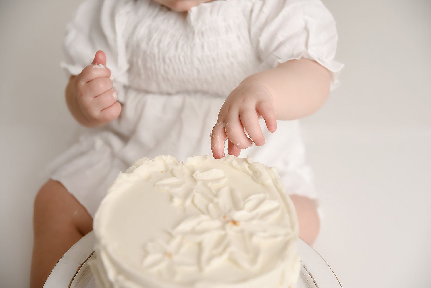 Close-up of baby girl in white dress reaching into a white frosted first birthday cake during cake smash session.