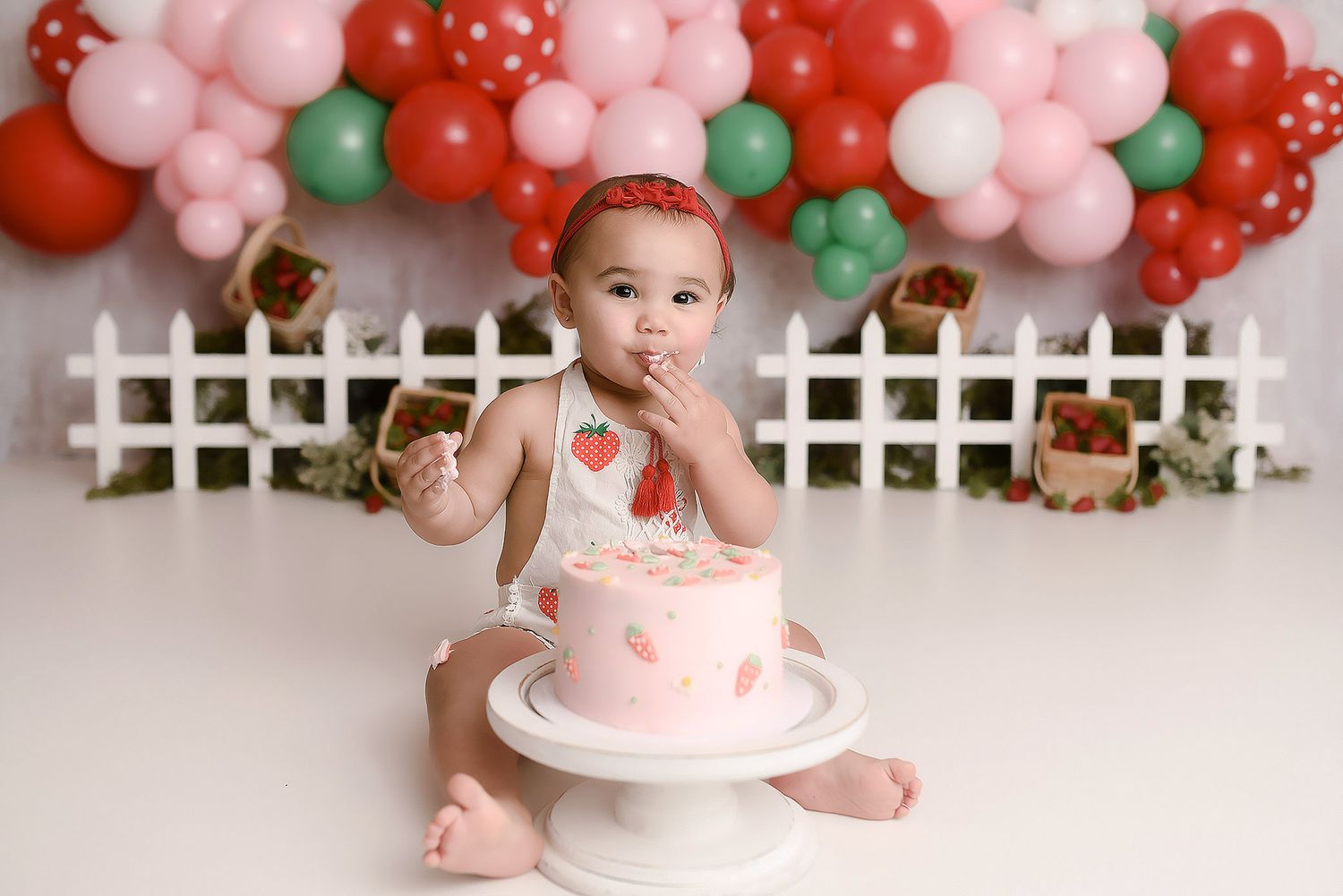 Baby girl eating pink strawberry smash cake with red and green balloon garland backdrop.