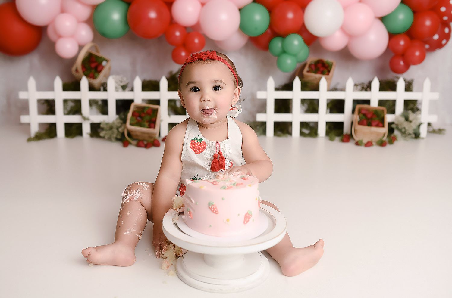 Baby girl in strawberry romper sitting beside pink smash cake with balloon backdrop.