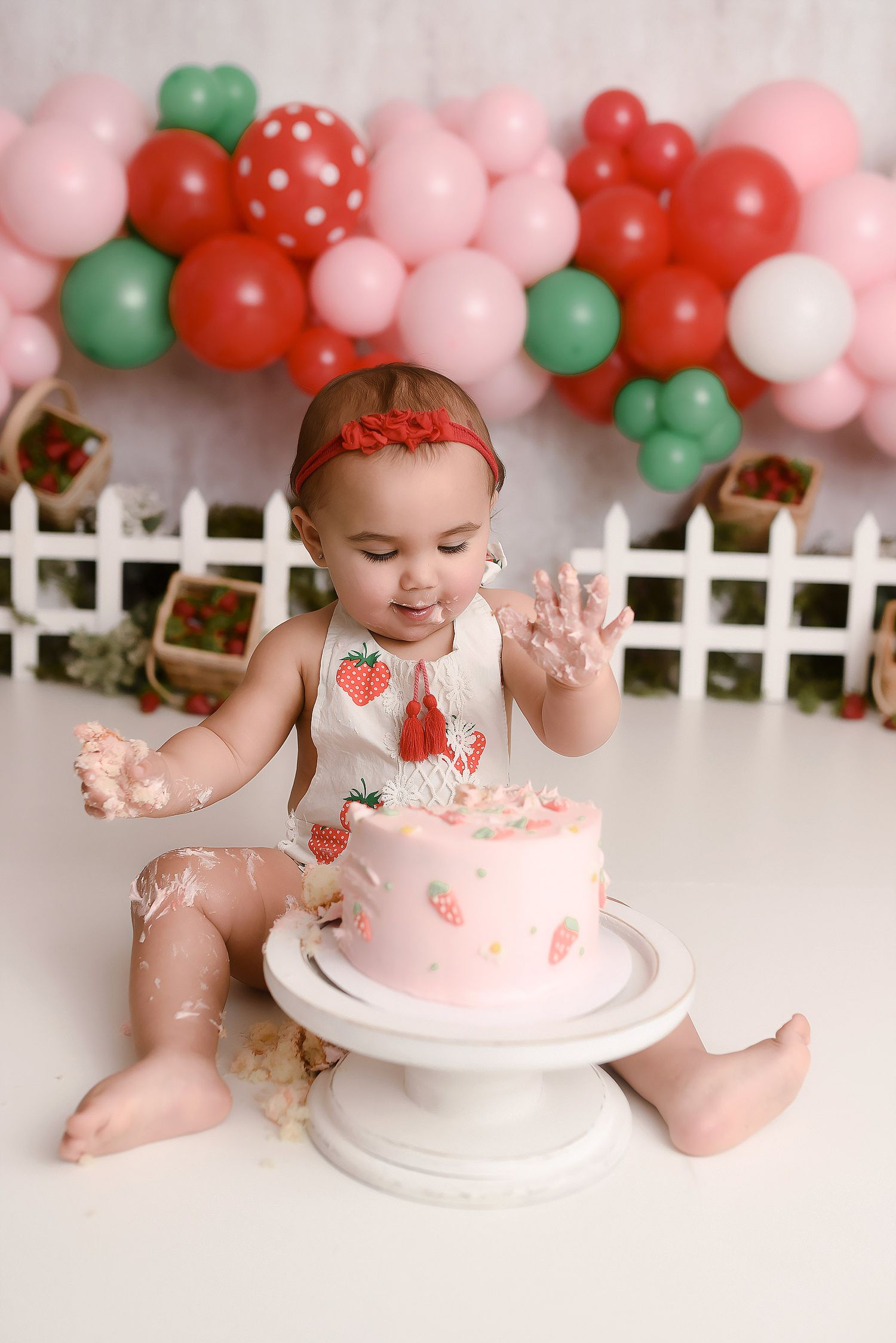 Baby girl in strawberry romper smashing pink birthday cake with balloon garland behind her.