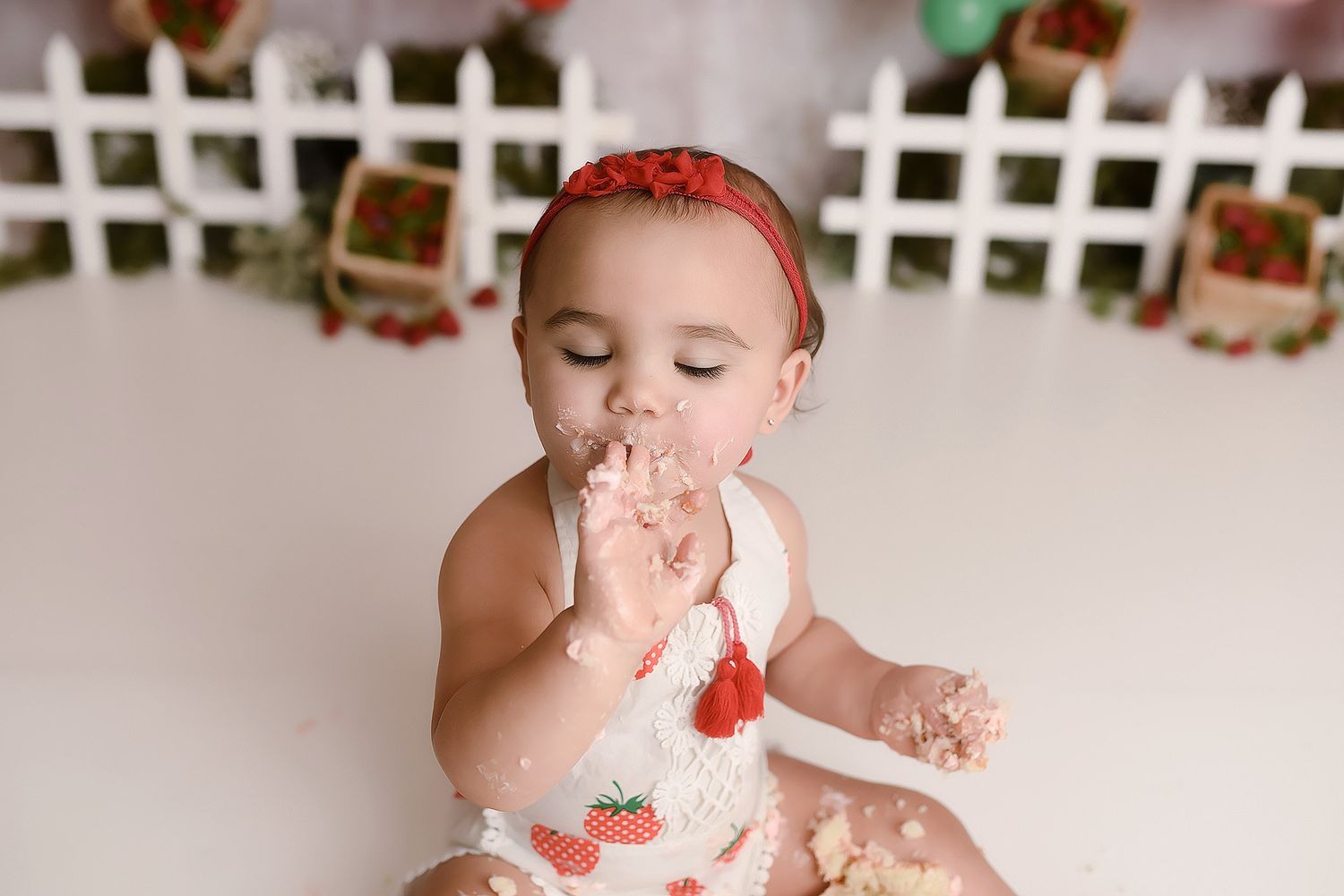Baby girl in strawberry outfit licking frosting off hands during cake smash session.