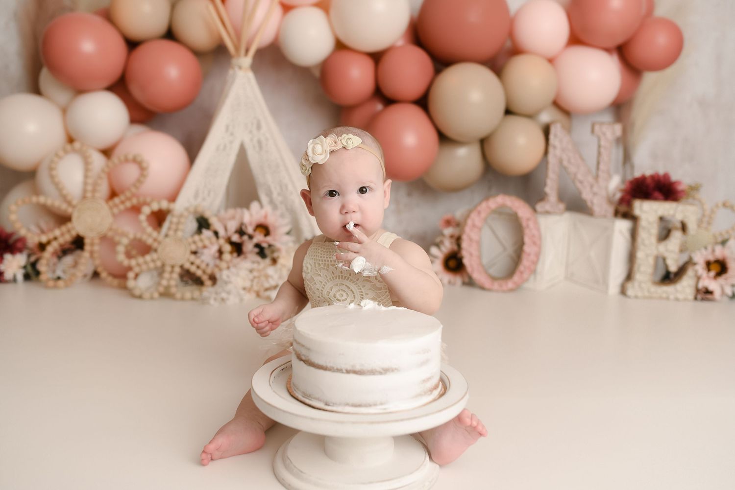 Baby girl eating white smash cake at boho teepee themed first birthday setup with balloon arch backdrop.