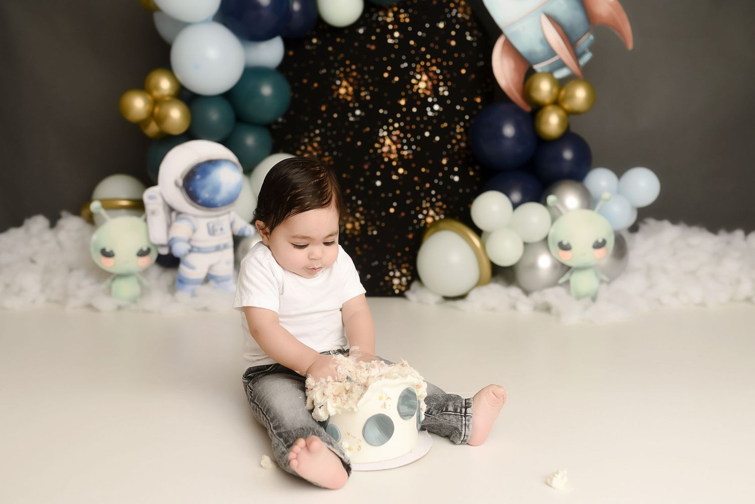 Baby boy in space-themed first birthday cake smash photo session with astronaut toys and balloon garland.