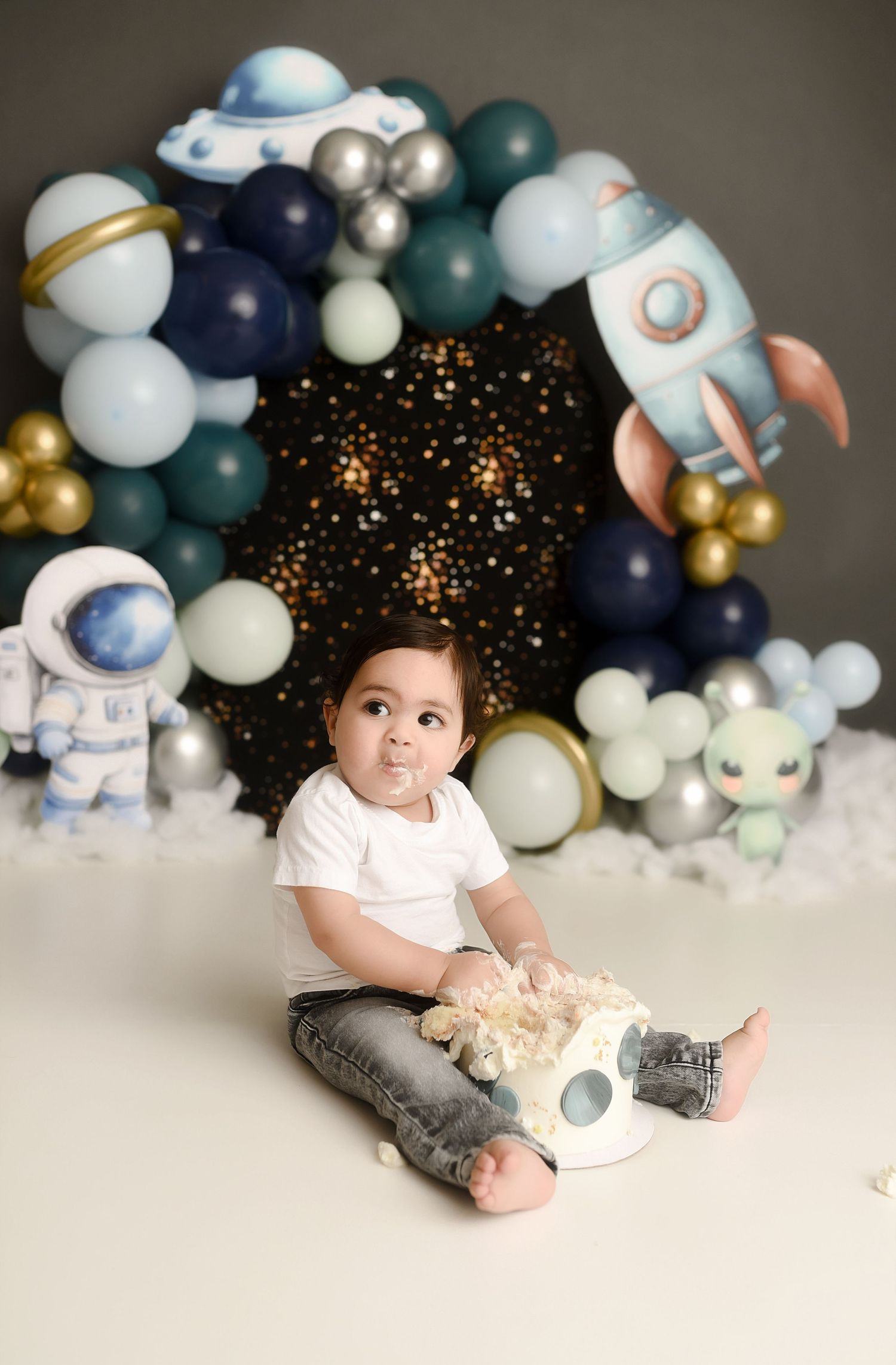 Baby boy sitting on floor with space-themed balloon arch, rocket, astronaut decor for a first birthday cake smash.