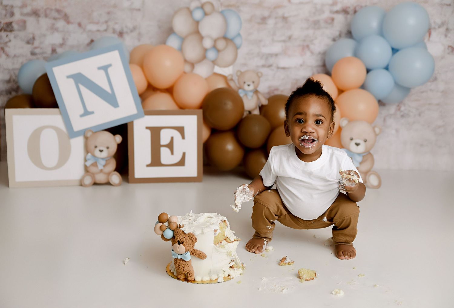Toddler boy smashing teddy bear cake with ONE blocks and blue balloon garland backdrop.