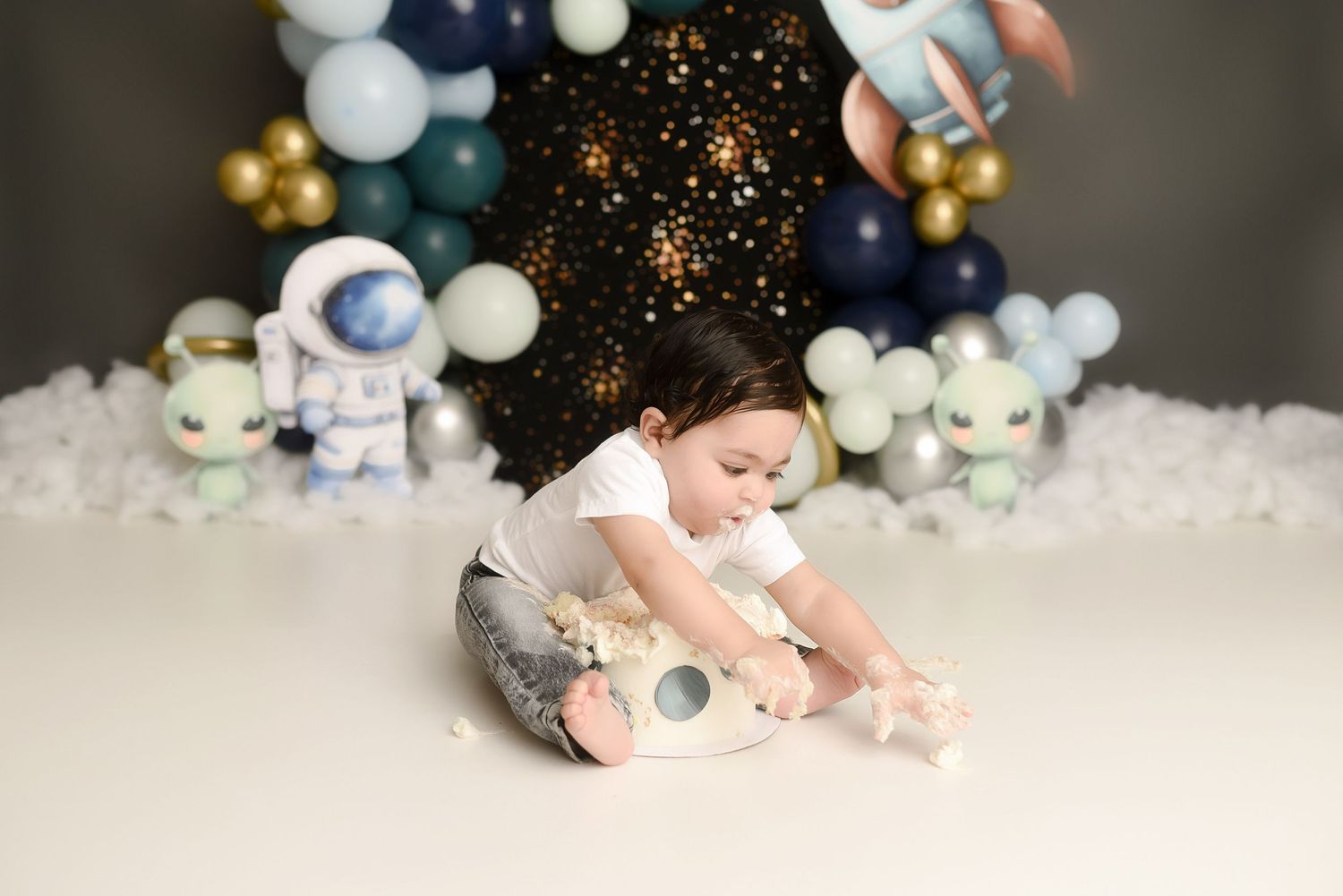 Child playing on floor with space-themed cake smash backdrop featuring astronaut toys and blue balloons.