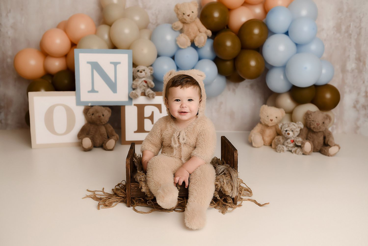 Baby boy in bear romper sitting in a wooden crate with ONE blocks and teddy bears for first birthday photos.