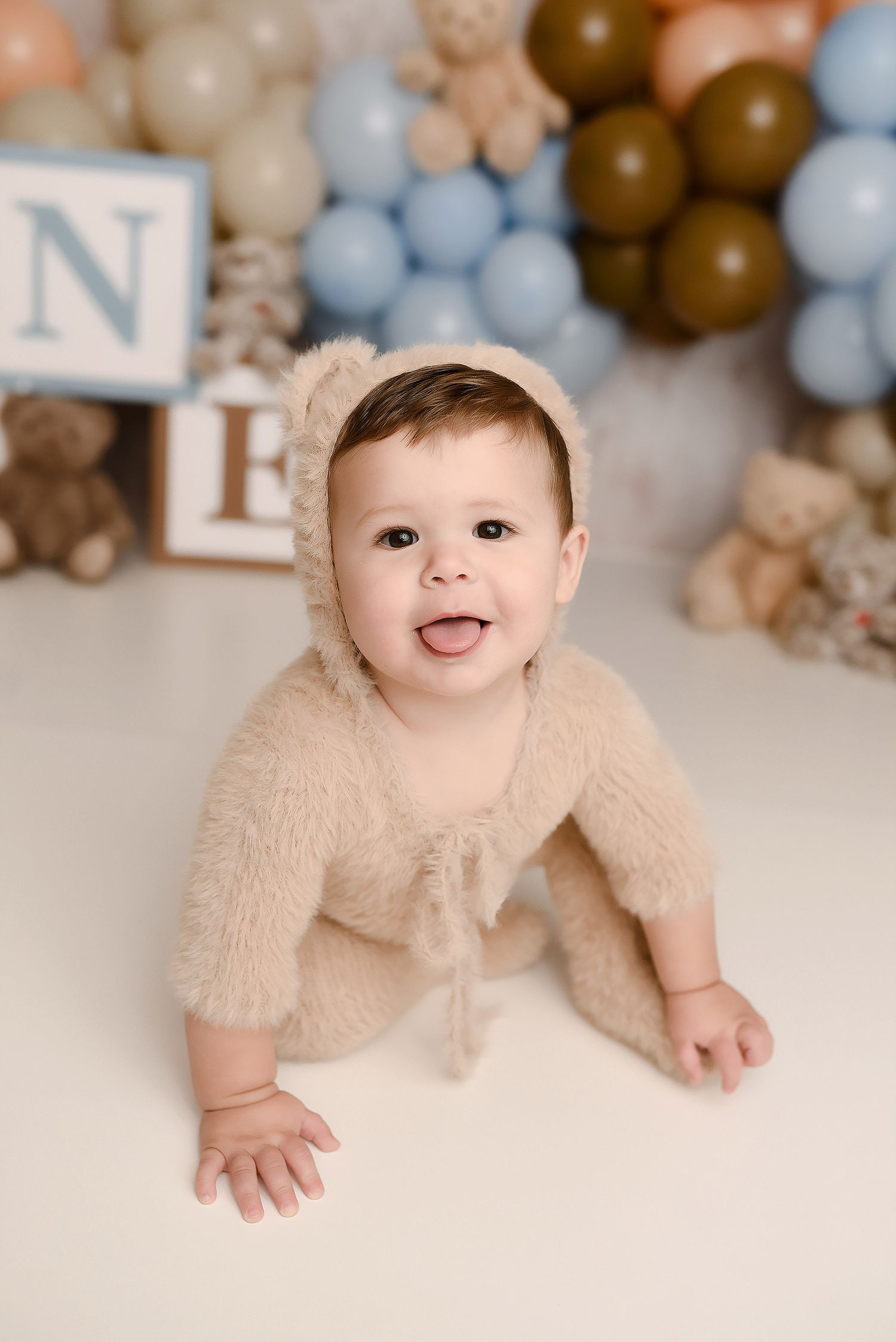 Smiling baby boy in fuzzy bear romper and bonnet crawling at a blue and brown balloon teddy bear birthday session.