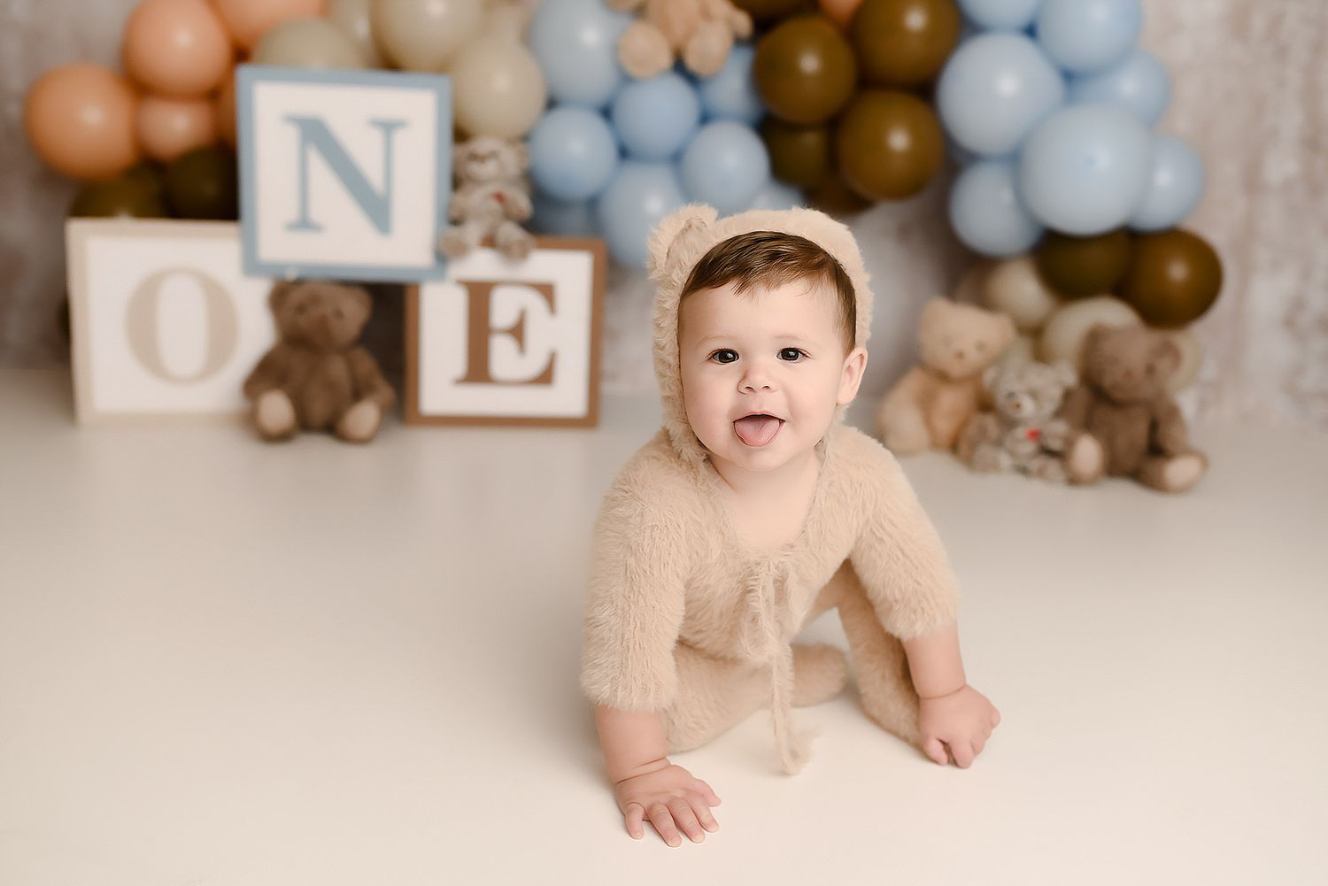 Cute baby boy in bear bonnet and romper smiling at a blue brown balloon ONE block teddy bear birthday session.