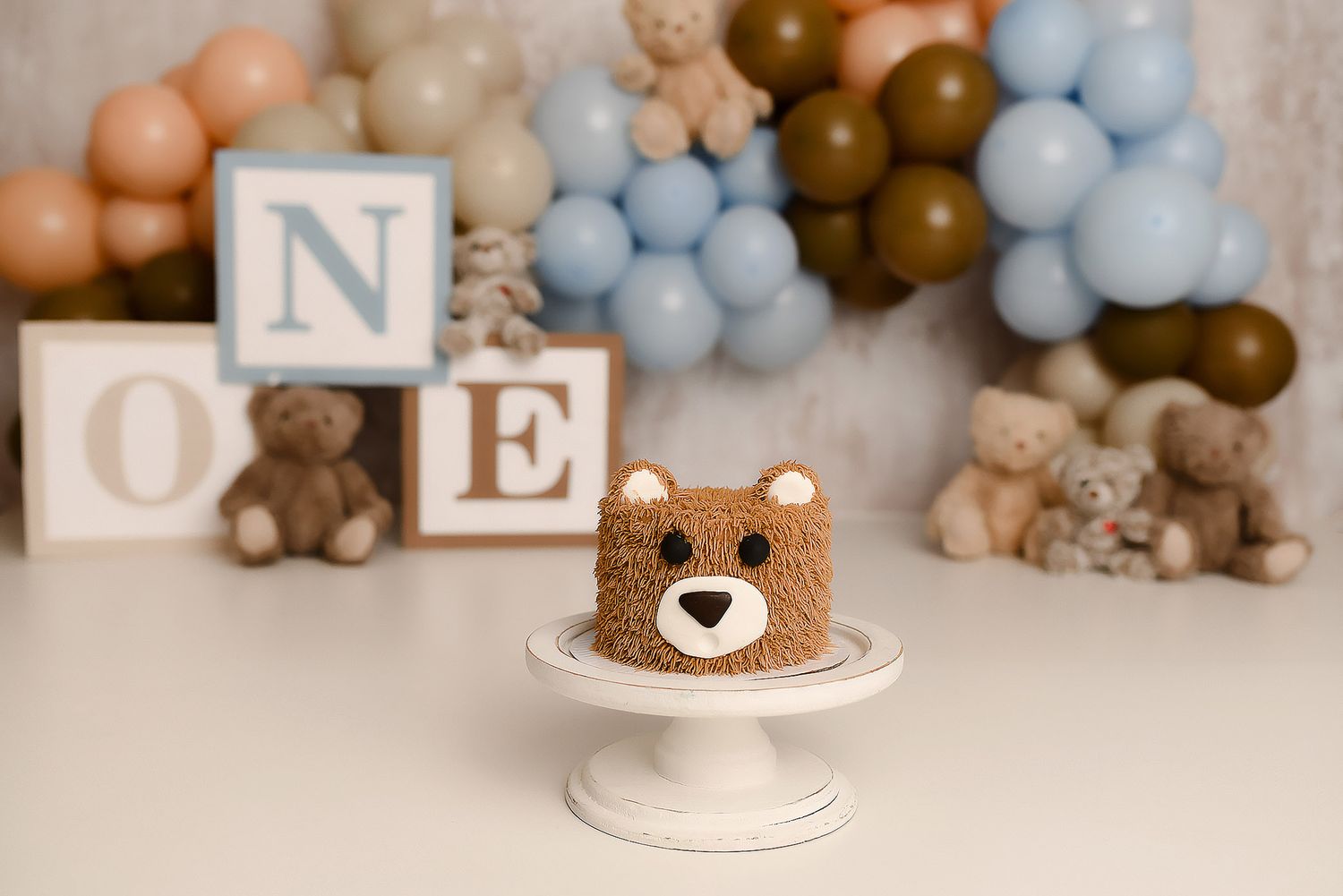 Teddy bear shaped brown cake on white stand with ONE blocks teddy bears and blue balloon backdrop.