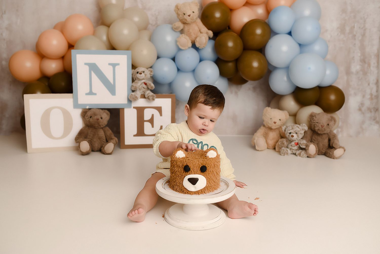 Baby boy in bear romper smashing a bear face cake at a teddy bear themed first birthday photo session.