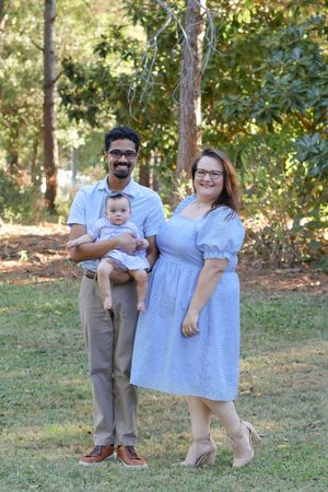 A family wearing blue and beige outfits poses together on grass in a wooded outdoor setting.
