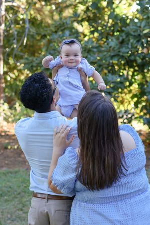 A family in white and grey clothing holds up a baby while enjoying time together outdoors on a sunny day.