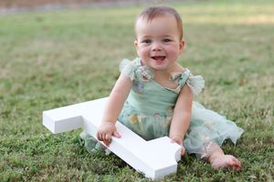 Baby in mint green dress sits on grass with white wooden number one prop during outdoor photo session.