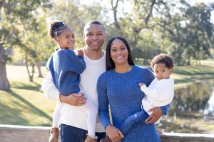Family wearing coordinating blue and white outfits poses together outdoors in a sunny park setting.