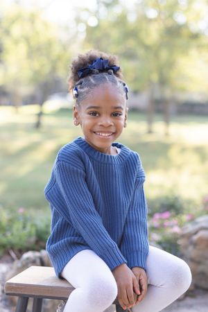 A child wearing a blue knit sweater and white pants sits outdoors with a bright smile on a sunny day.