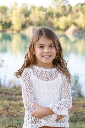 A child in a white lace dress poses outdoors by a lake on an autumn day.