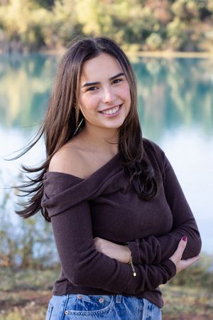 A person in a brown sweater and jeans poses outdoors by a lake on a sunny autumn day.
