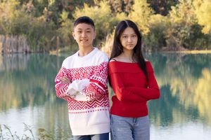 Couple in winter sweaters poses by tranquil lake surrounded by autumn foliage and mountains.