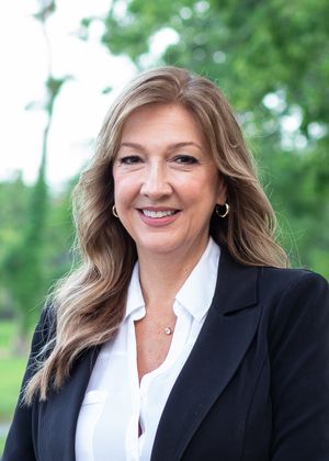 Professional wearing a black blazer and white blouse smiles against a natural outdoor backdrop with green foliage.