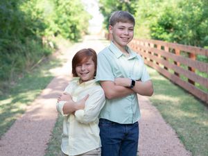 Two young siblings stand back-to-back on a garden path surrounded by greenery and a wooden fence.