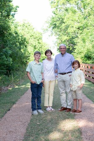 A family group stands together on a brown gravel path surrounded by green trees in a park setting.