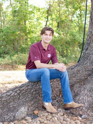 Casual outdoor portrait showing someone in a maroon polo and jeans sitting on a large tree trunk in a wooded setting.