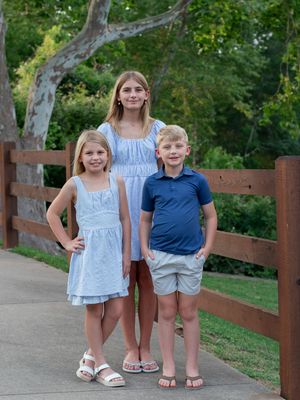 Three young siblings stand together on a path with a wooden fence and lush greenery in the background.