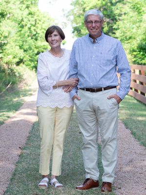 A couple stands together on a gravel path in a garden setting wearing casual summer attire.