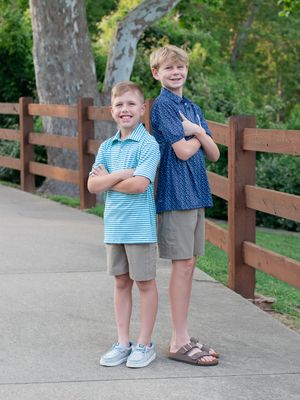 Two young children stand together on a path with a wooden fence and trees in the background.
