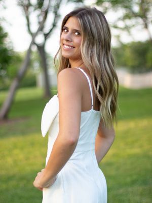 A person in a white dress poses gracefully in a grassy outdoor setting with trees in the background.