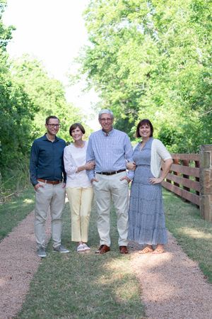 Four people in casual summer attire stand together on a gravel path surrounded by green trees and a wooden fence.