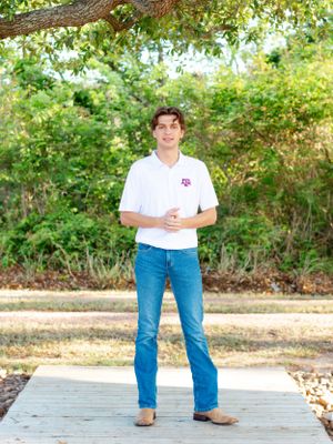 Person in white polo shirt and blue jeans standing on wooden deck in front of lush garden background.