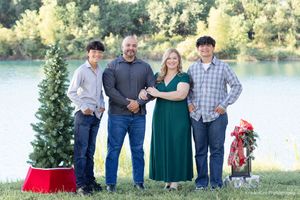 A family stands together outdoors by a lake with a decorated Christmas tree and festive holiday decor.