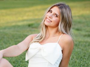 Outdoor portrait of someone wearing a white sundress smiling while sitting on grass on a sunny day.