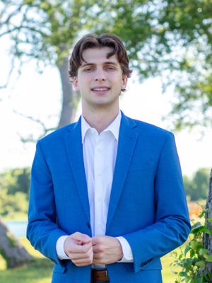 A person wearing a bright blue suit jacket and white dress shirt stands outdoors on a sunny day with trees in the background.