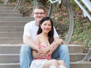 A couple sits together on outdoor concrete steps with decorative metal railing on a sunny day.