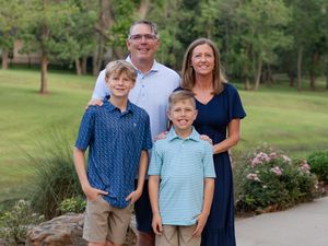 A family of four poses outdoors in front of landscaping and trees on a summer day.