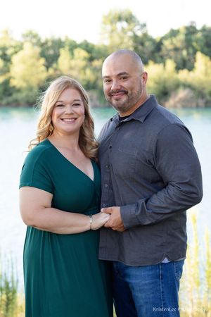A couple in a green dress and gray shirt poses together by a lakeside during an outdoor photo session.