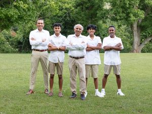 A wedding party wearing white shirts and khaki pants stand casually on lush green grass outdoors.