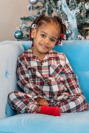 Child wearing plaid pajamas sitting on blue couch next to decorated Christmas tree.