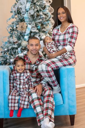 Family wearing matching plaid pajamas poses together on blue couch by Christmas tree.