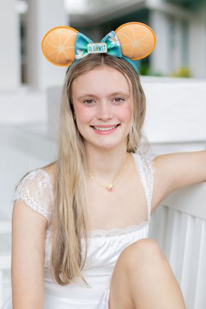 A person wearing a white dress and Mickey Mouse ears with a blue bow poses outdoors on a sunny day.